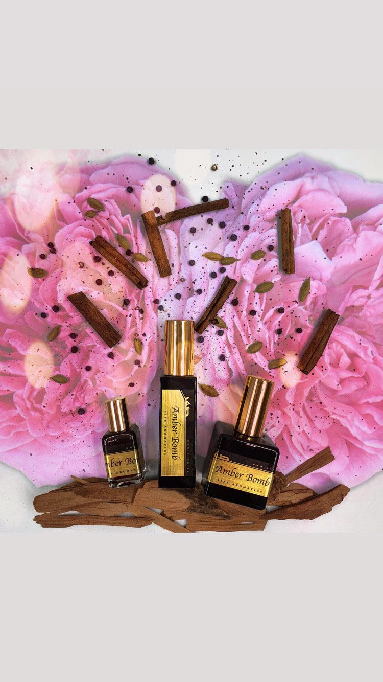 Amber Bomb by Sifr Aromatics in 15ml oil roller ball and 50ml Eau De Parfum spray bottles. Surrounded by exotic oriental spices like cardamom, cinnamon, and sandalwood. A large rose blooms in the background, enhancing the rich rose and amber scent.