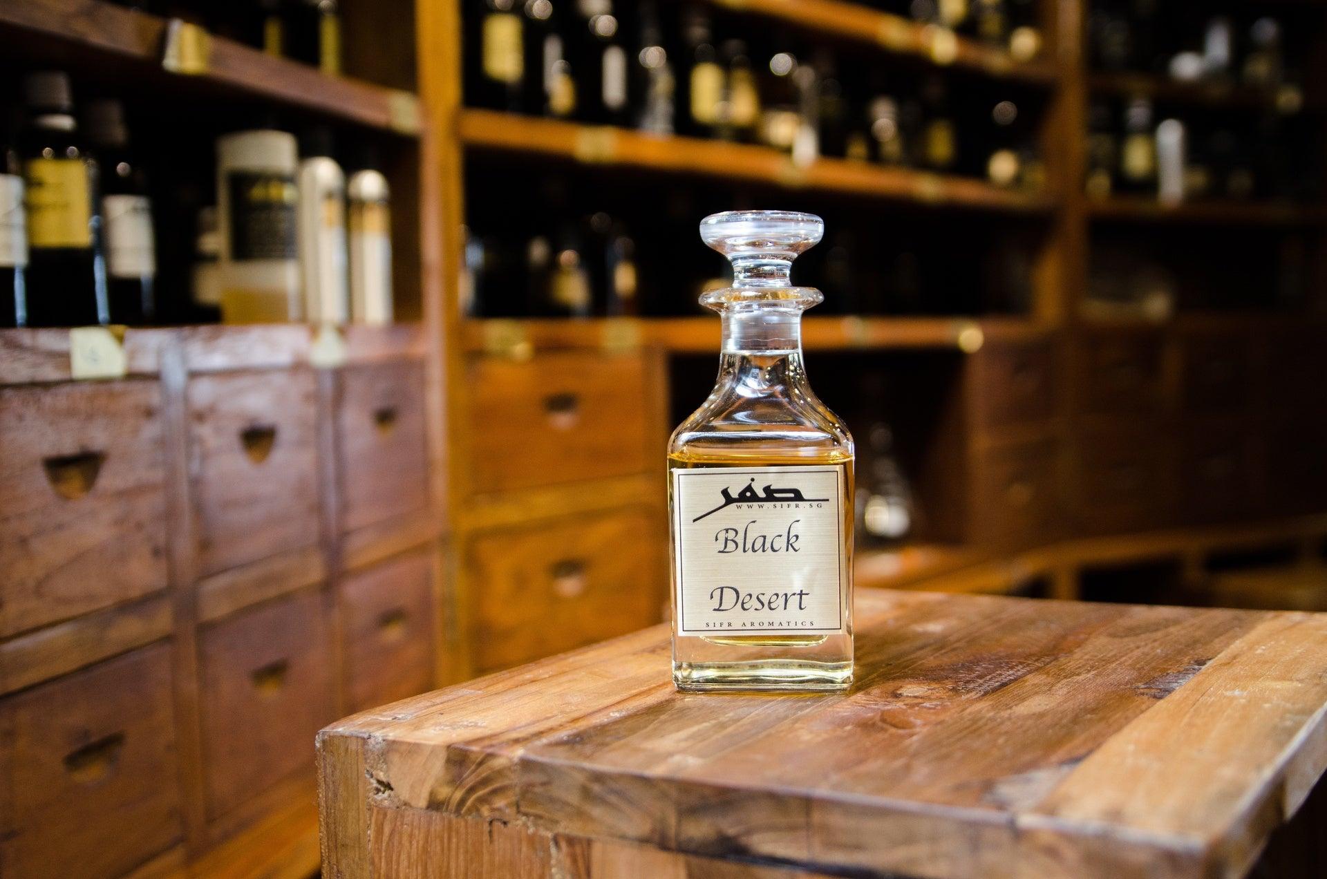 Ornate glass bottle for perfume oil with a gold label displaying the Sifr Aromatics logo and the name 'Black Desert.' The background features an out-of-focus wooden perfume organ with various ingredients, creating an aesthetically pleasing scene