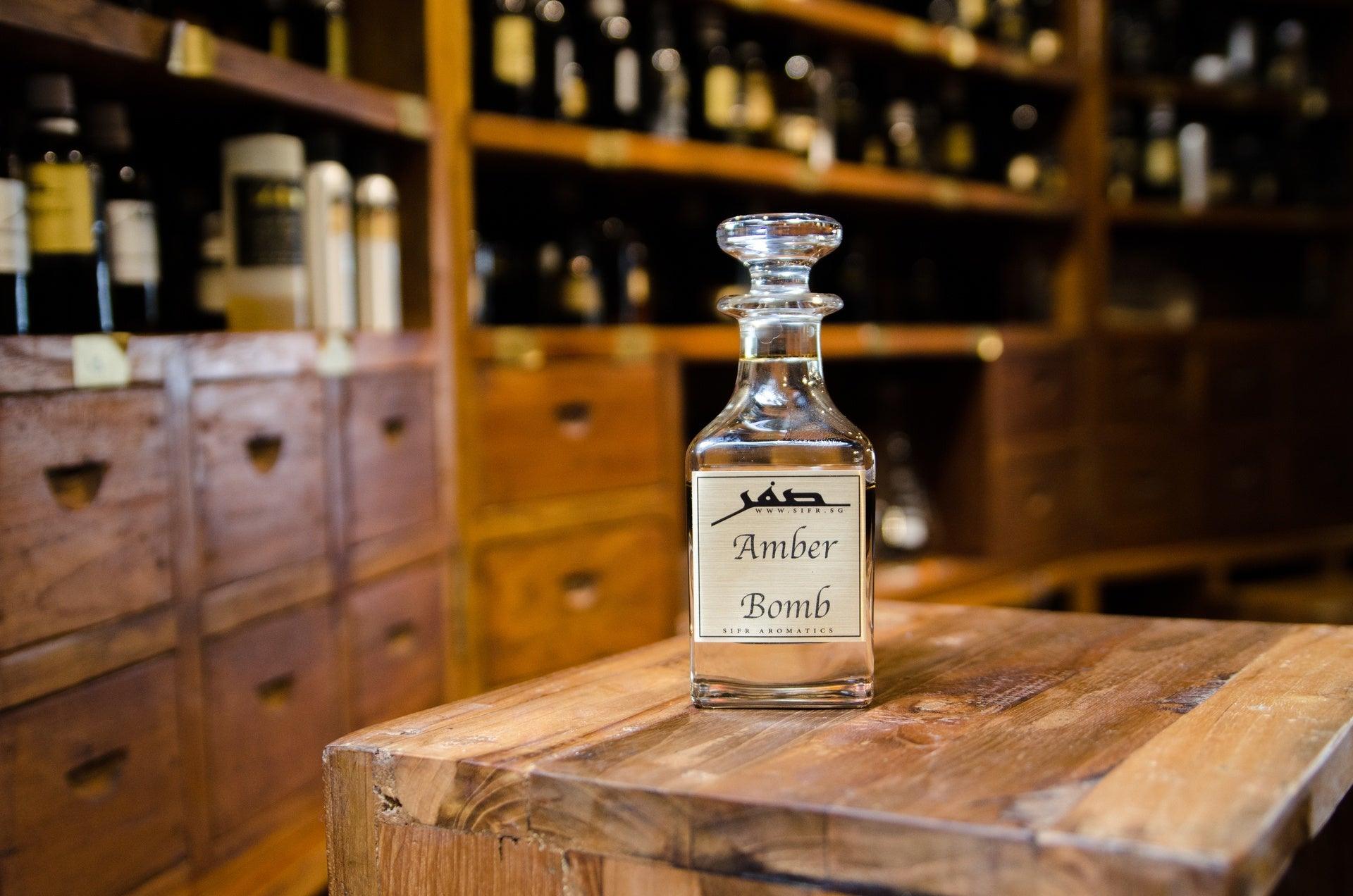 Ornate glass bottle for perfume oil with a gold label displaying the Sifr Aromatics logo and the name 'Amber Bomb.' The background features an out-of-focus wooden perfume organ with various ingredients, creating an aesthetically pleasing scene.